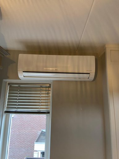 Een wandairconditioner gemonteerd op een lichte muur met een venster hieronder.